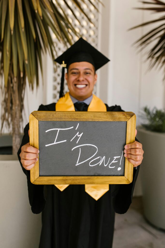 Smiling graduate in cap and gown holding 'I'm Done!' sign outdoors.